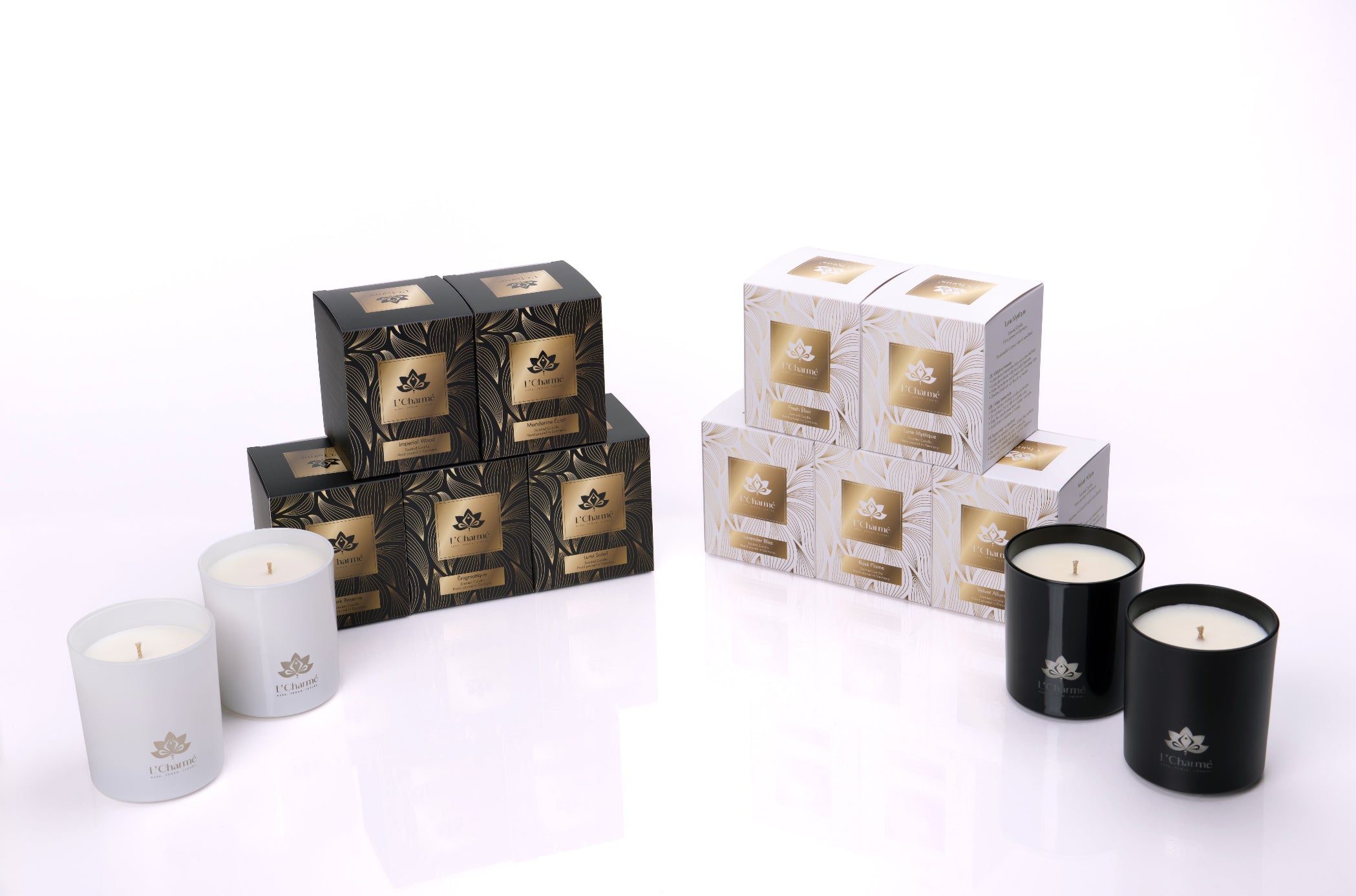 L’Charmé Duftkerzen Sortiment mit kleinen Kerzen in weißem und schwarzem Glas vor gestapelten Geschenkboxen in Schwarz/Gold und Weiß/Gold, Produktfoto auf weißem Hintergrund