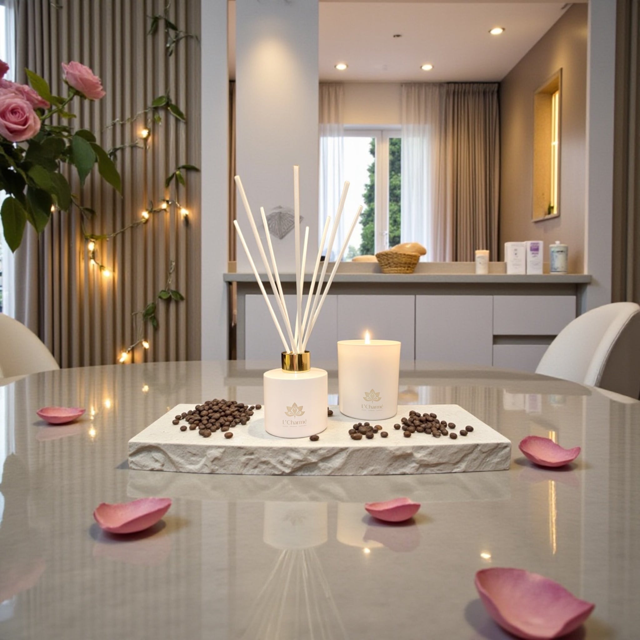 Weißer Duftdiffuser und brennende Duftkerze von L’Charmé auf einer Steinplatte mit Kaffeebohnen und rosa Blütenblättern im eleganten Wohnraum