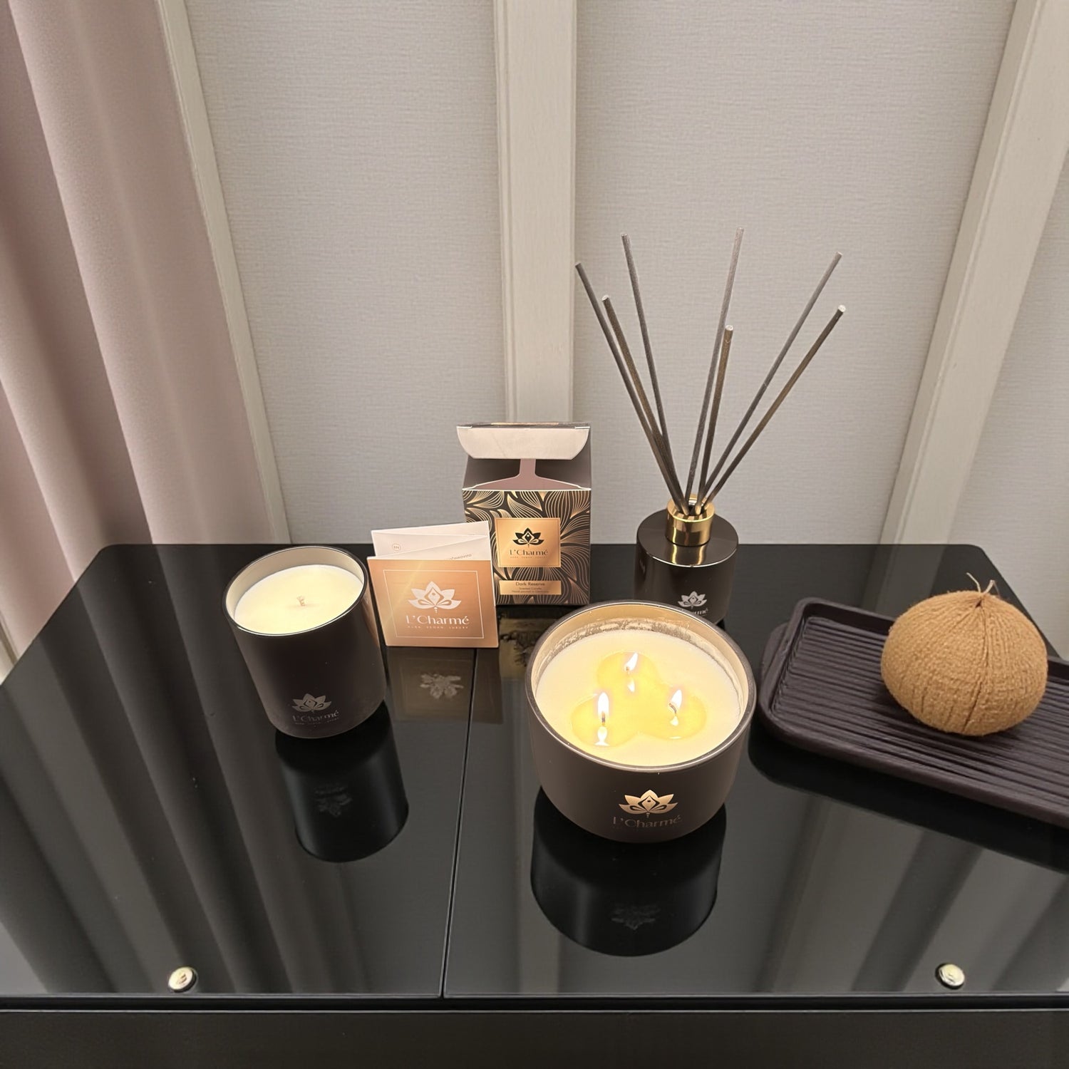 Duftkerzen und Duftstäbchen von L’Charmé mit Verpackung auf einem schwarzen Glastisch in elegantem Arrangement