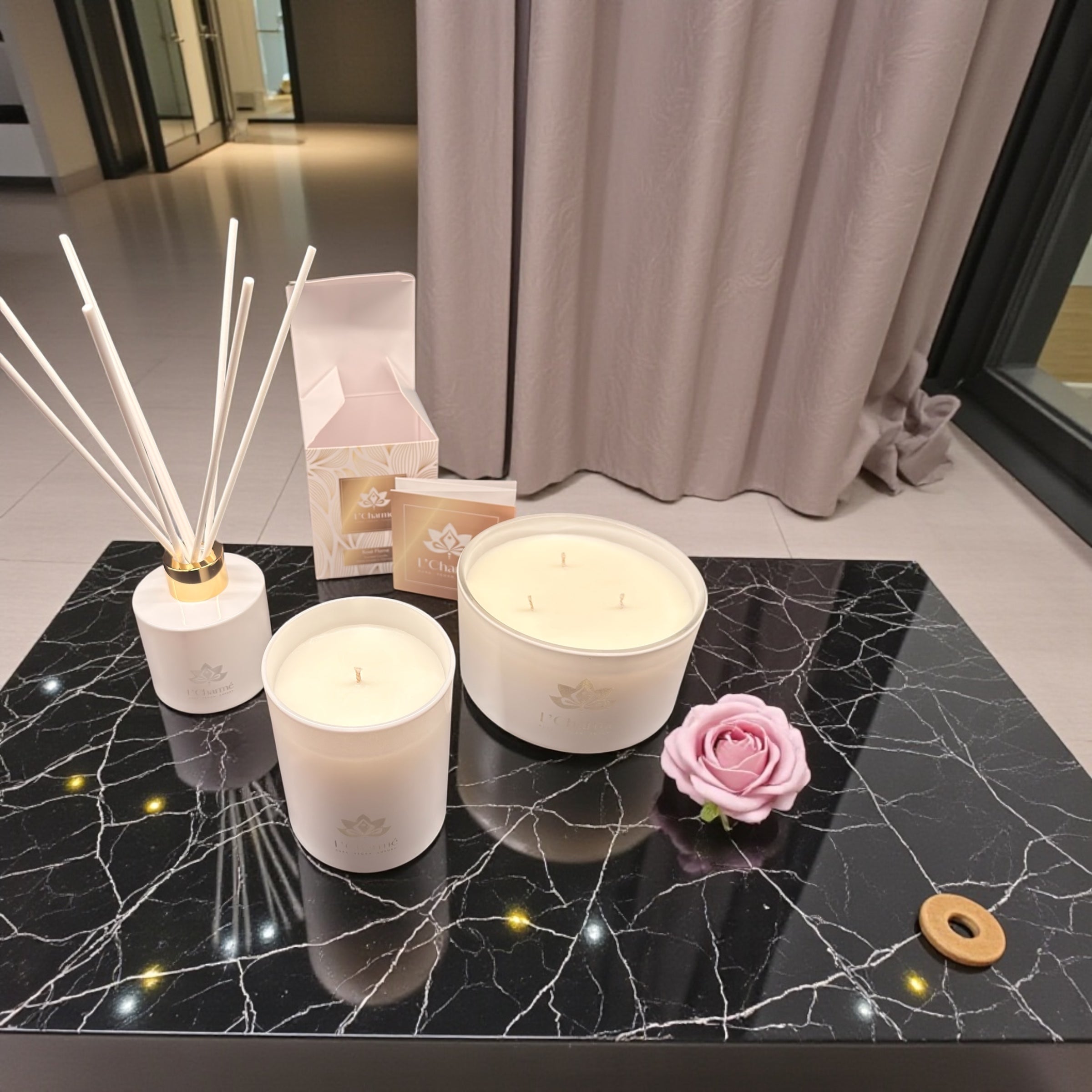 Duftkerzen und Duftstäbchen von L’Charmé mit Verpackung auf einem schwarzen Marmortisch mit rosa Rose in elegantem Ambiente