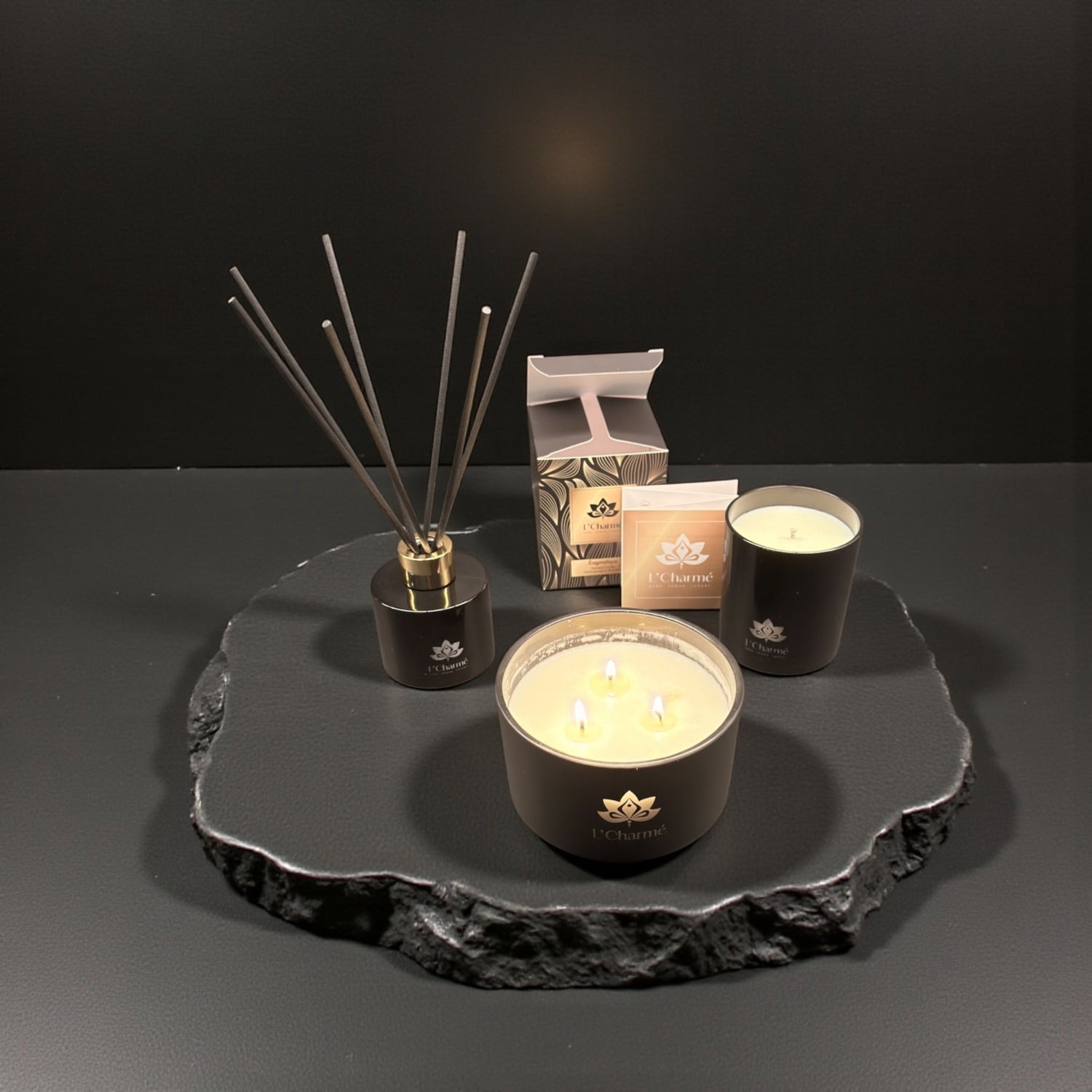 Duftkerzen und Duftstäbchen von L’Charmé mit Verpackung auf einer schwarzen Natursteinplatte in elegantem Arrangement