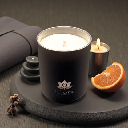 Schwarze Duftkerze von L’Charmé auf einer runden Steinplatte mit Orange, Spa-Steinen und brennendem Teelicht in elegantem Ambiente