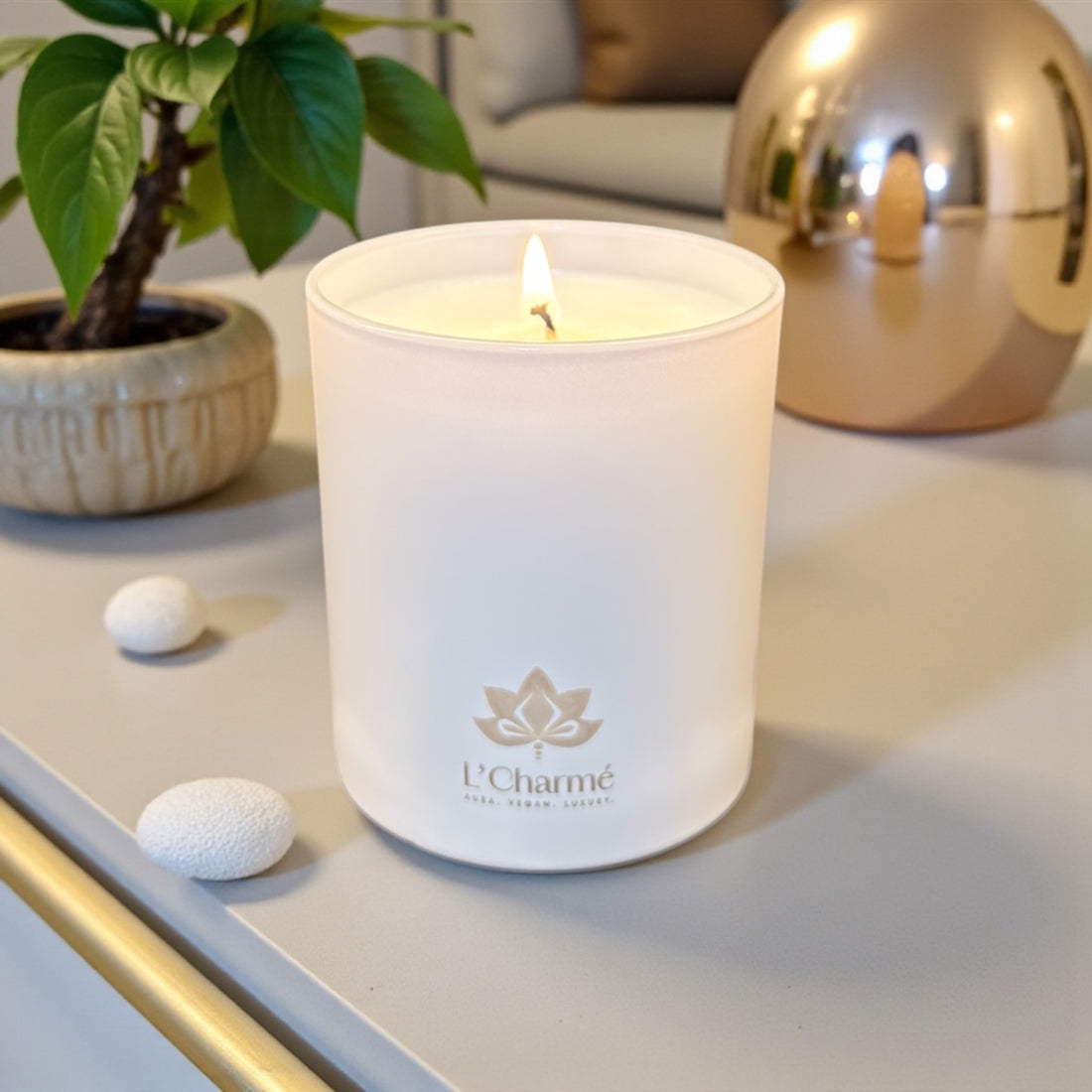 Weiße brennende Duftkerze von L’Charmé auf einem hellen Tisch neben einer Pflanze und goldener Dekoration in elegantem Ambiente