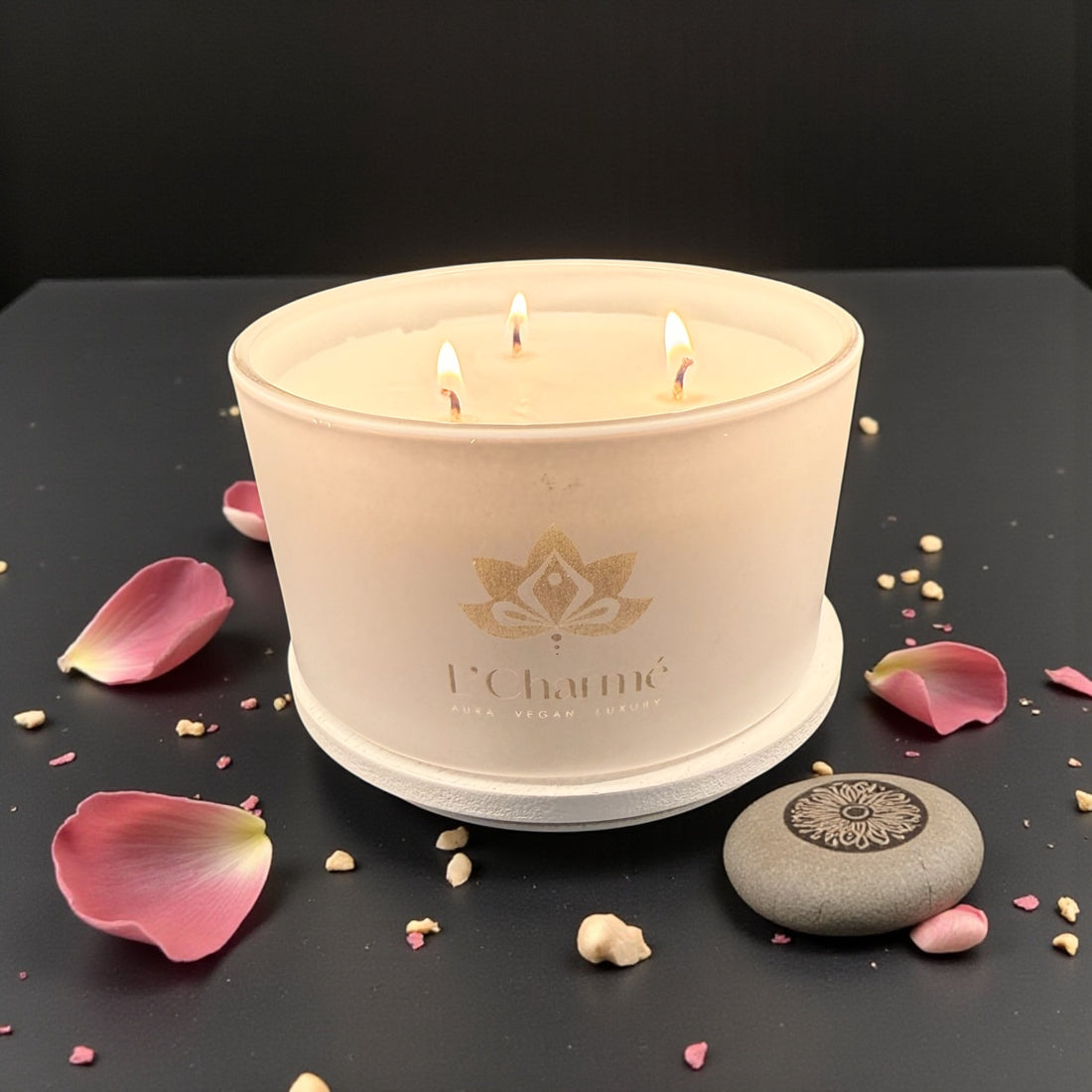 Weiße Dreidocht-Duftkerze von L’Charmé mit brennenden Flammen auf einem schwarzen Tisch mit rosa Rosenblättern in elegantem Ambiente