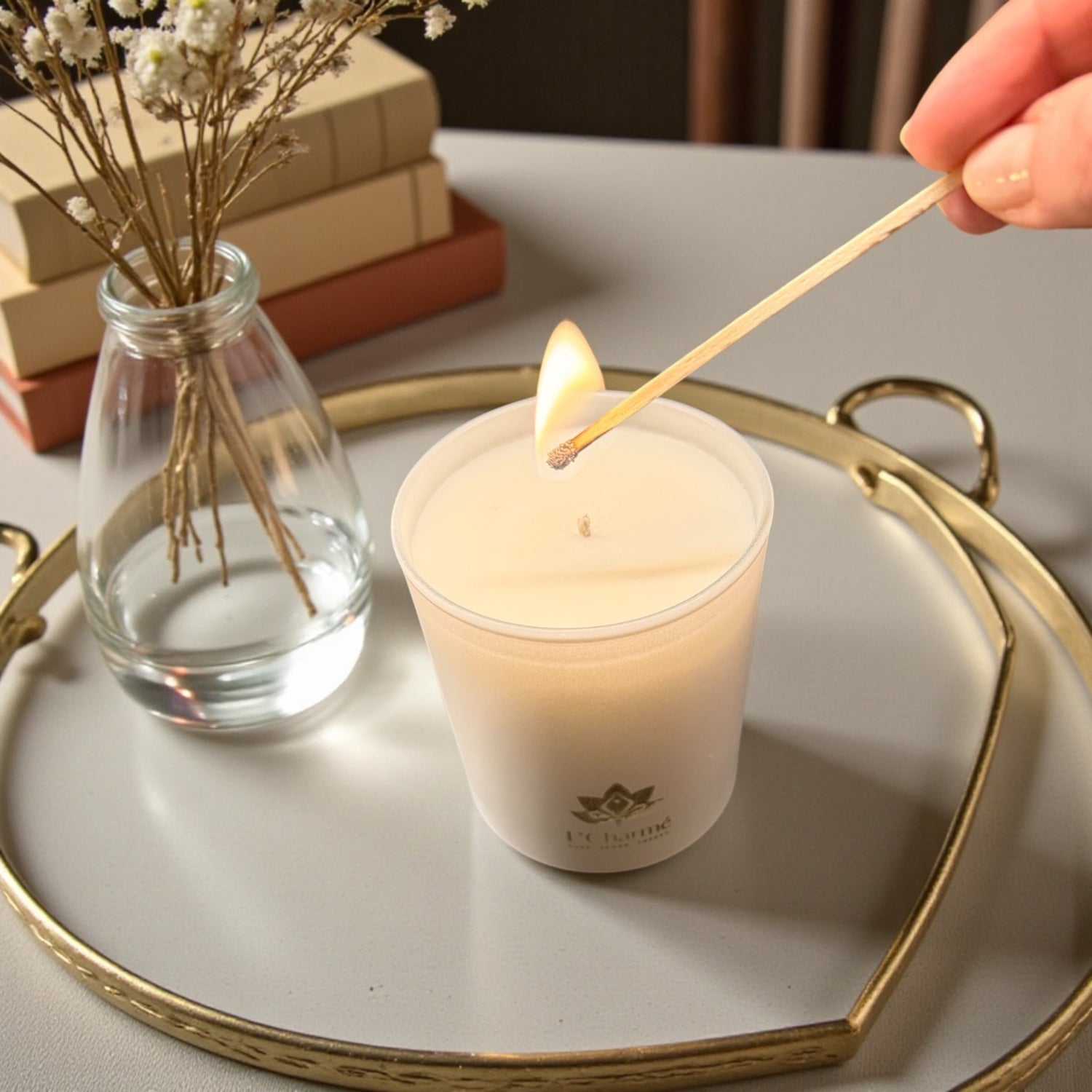 Weiße Duftkerze von L’Charmé auf einem goldenen Tablett, während sie mit einem Streichholz angezündet wird, neben einer Glasvase mit Trockenblumen