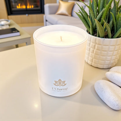 Weiße Duftkerze von L’Charmé auf einem hellen Tisch neben einer Pflanze und weißen Steinen in elegantem Wohnambiente