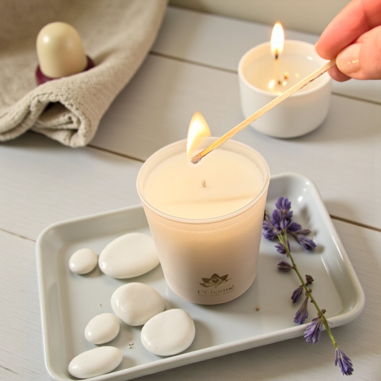 Weiße brennende Duftkerze von L’Charmé auf einem weißen Tablett mit Lavendel und weißen Steinen, während sie angezündet wird