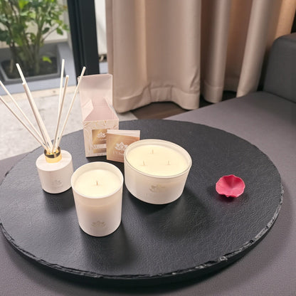 Weiße Duftkerzen und Duftstäbchen von L’Charmé mit Verpackung auf einer runden schwarzen Platte mit rosa Rosenblatt in elegantem Wohnambiente