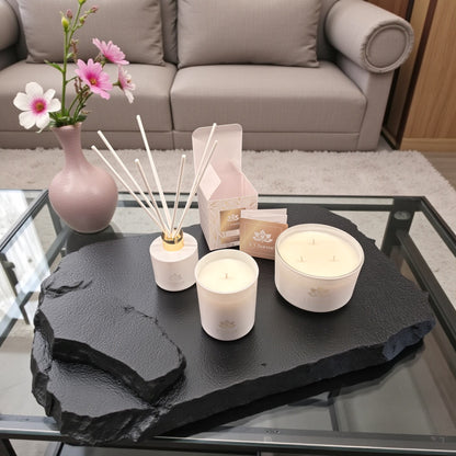 Weiße Duftkerzen und Duftstäbchen von L’Charmé mit Verpackung auf einer schwarzen Natursteinplatte neben einer rosa Blumenvase in elegantem Wohnambiente
